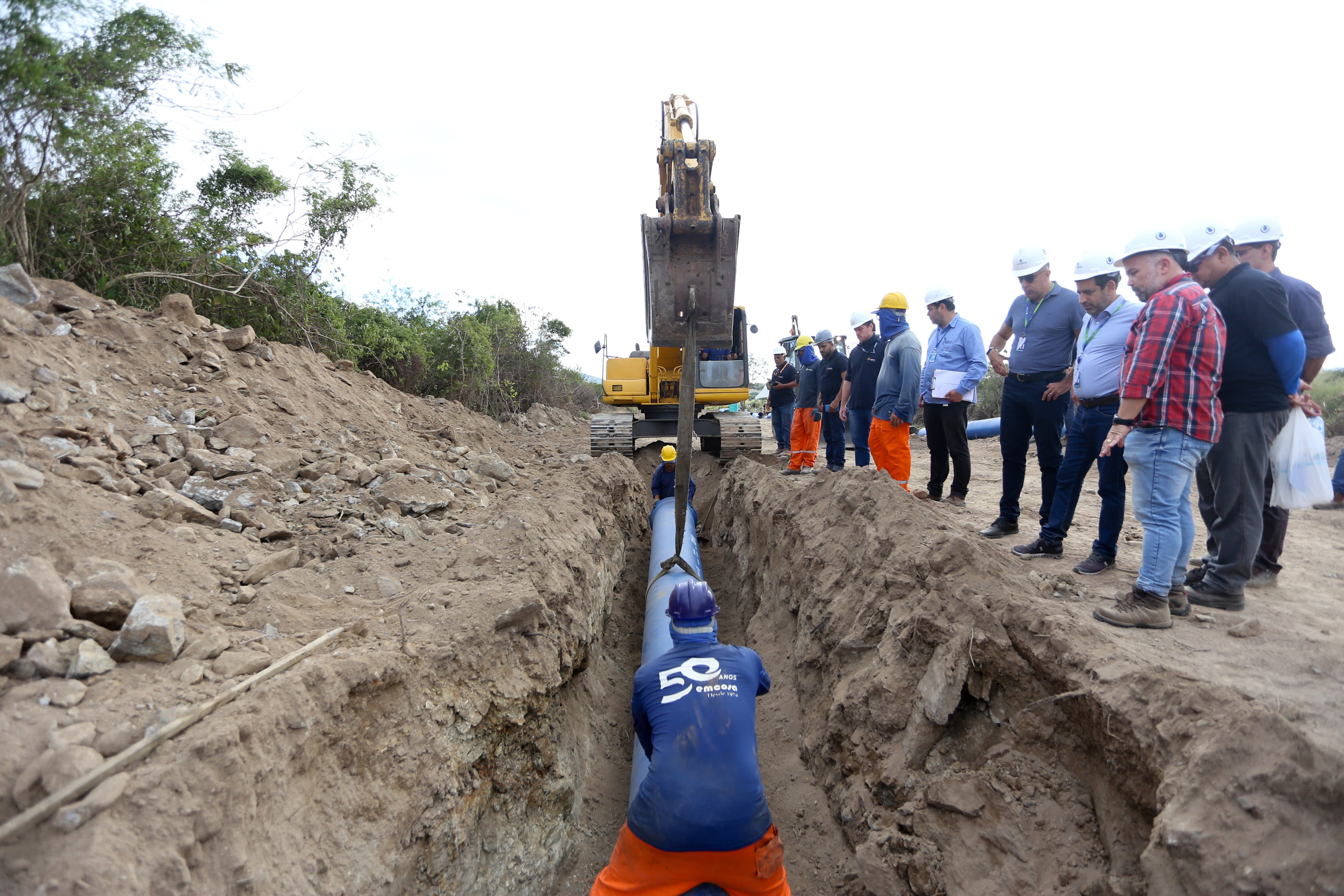 Foto da obra da Adutora do Agreste em Bezerros-PE. A imagem apresenta máquinas e homens trabalhando em uma vala onde está sendo inserido um cano para abastecimento de água. Há diversos homens acima da vala observando o trabalho realizado.